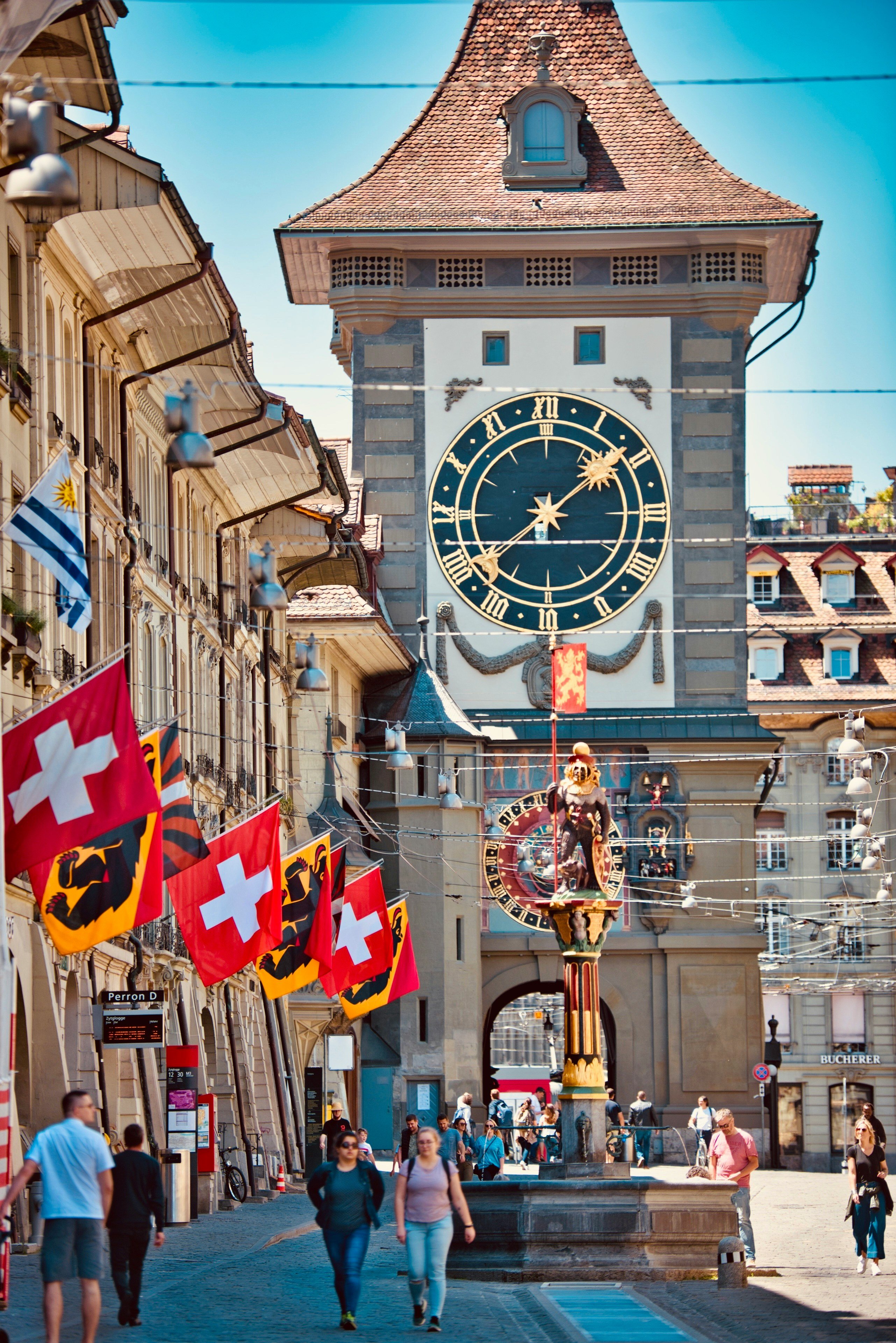 Bern’s Zytglogge clock tower with Swiss flags along the street and people walking in the historic city center.
